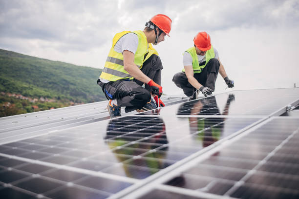 Two people, male engineers with protective helmets installing photovoltaic panel system. Professional electricians working on a roof.